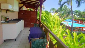 A balcony with a table and chairs, a kitchen area with a microwave, and a view of a pool and palm trees. - Emerald b (Sainte-Rose)