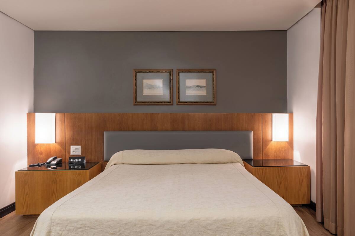 A hotel room with a large bed, wooden headboard, two bedside tables with lamps, a telephone, and a desk with a laptop.