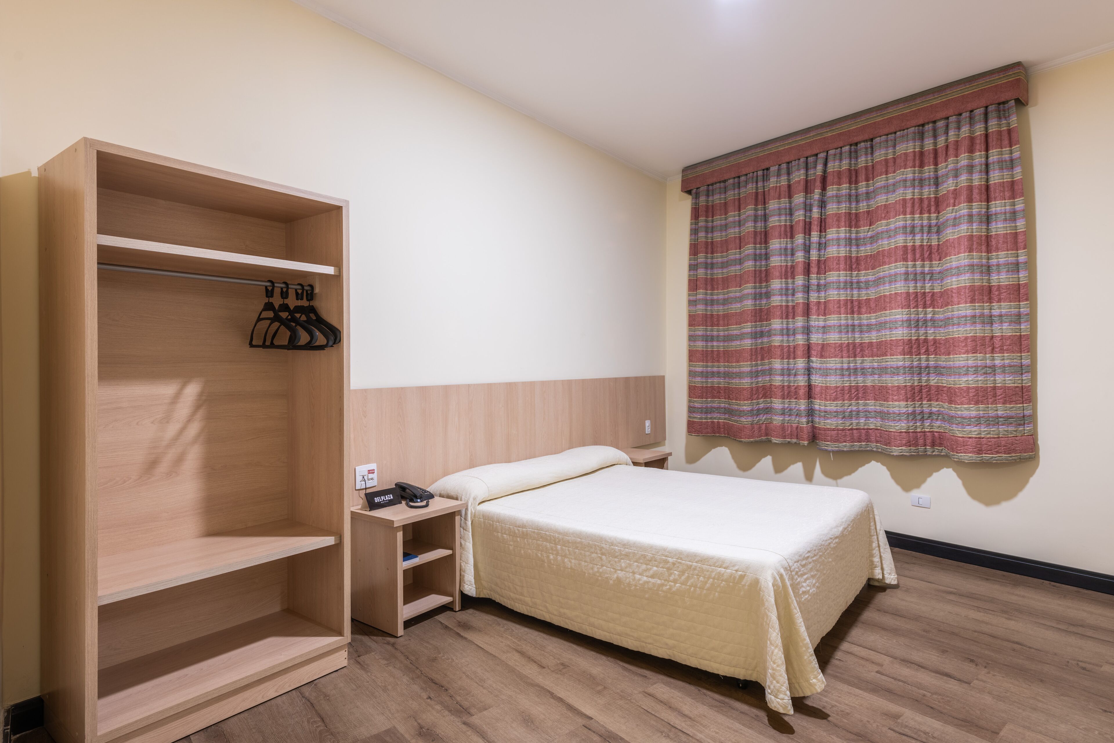 Ein Schlafzimmer mit einem Bett, einem Holzschrank mit Kleiderhaken, einem Nachttisch und einem Fenster mit Vorhängen.