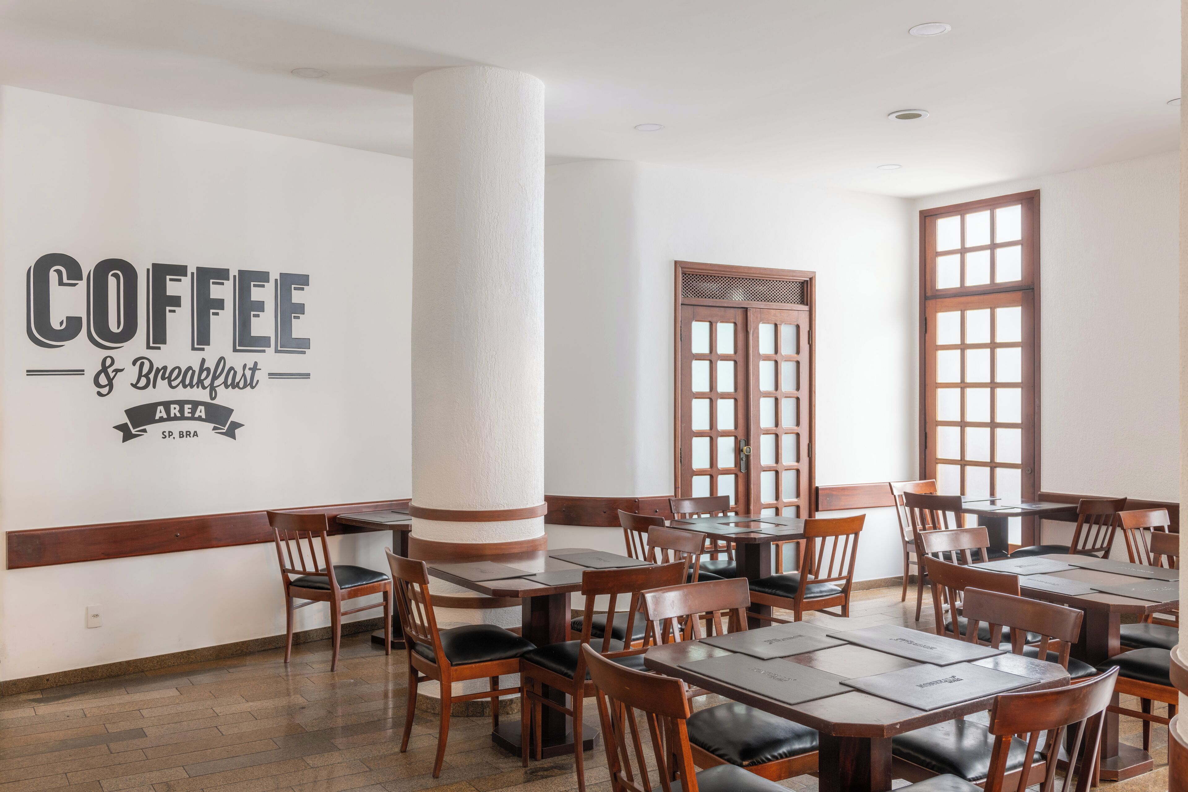Un restaurante con varias mesas y sillas de madera, un grueso pilar blanco, y un letrero en la pared que dice "COFFEE & Breakfast AREA".