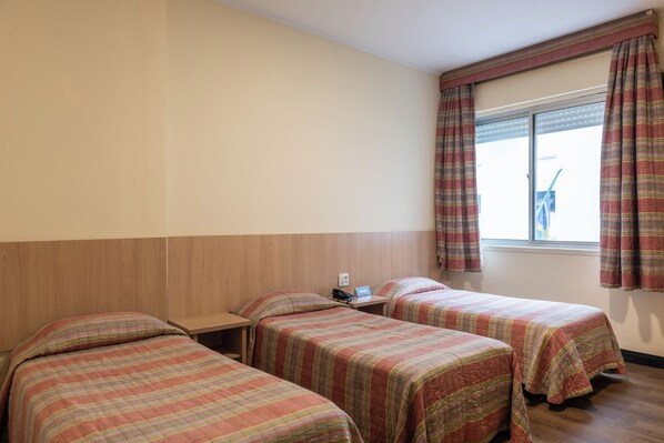 A hotel room with three beds, a wooden headboard, a window with curtains, and a small table with a phone and a sign.