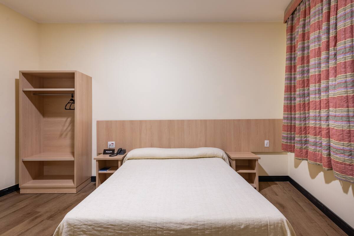 A hotel room with a bed, a wooden wardrobe, a bedside table with a phone, and a striped curtain.