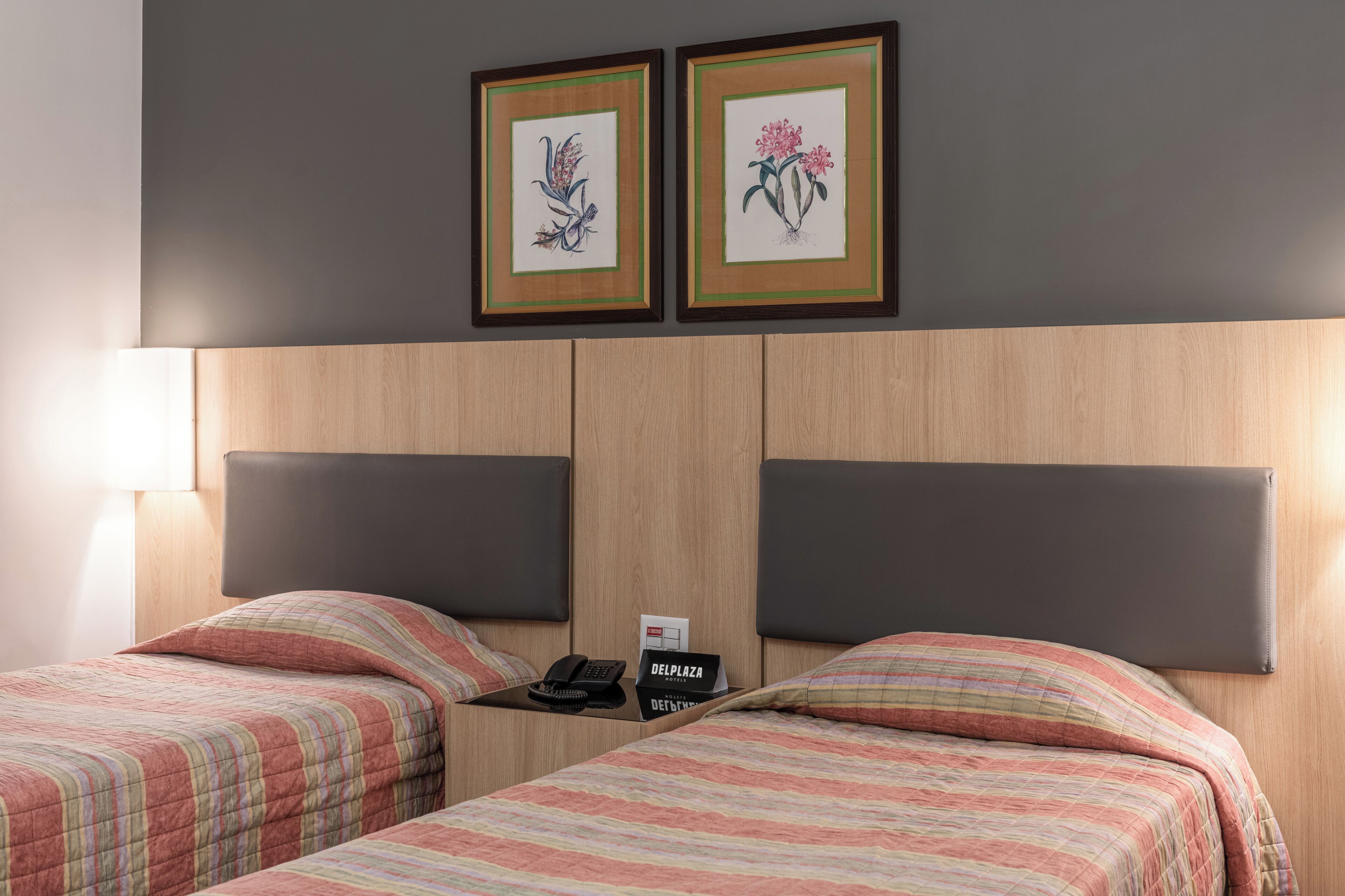 Une chambre d’hôtel avec deux lits, une table de chevet avec un téléphone et un panneau, et deux tableaux encadrés au mur.