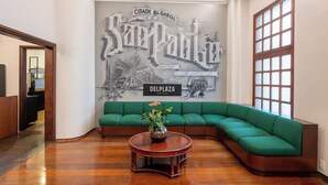 A lobby with a green sofa, a round wooden coffee table with a flower vase, and a large wall mural. - DELPLAZA Timbiras São Paulo (São Paulo)