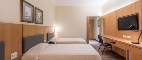 Quarto de hotel com duas camas, uma escrivaninha, uma cadeira, uma TV e quadros nas paredes.
