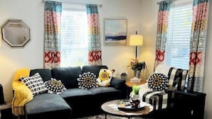 A living room with a sofa, ottoman, coffee table, and a rug. There is a lamp, a plant, and a painting on the wall.