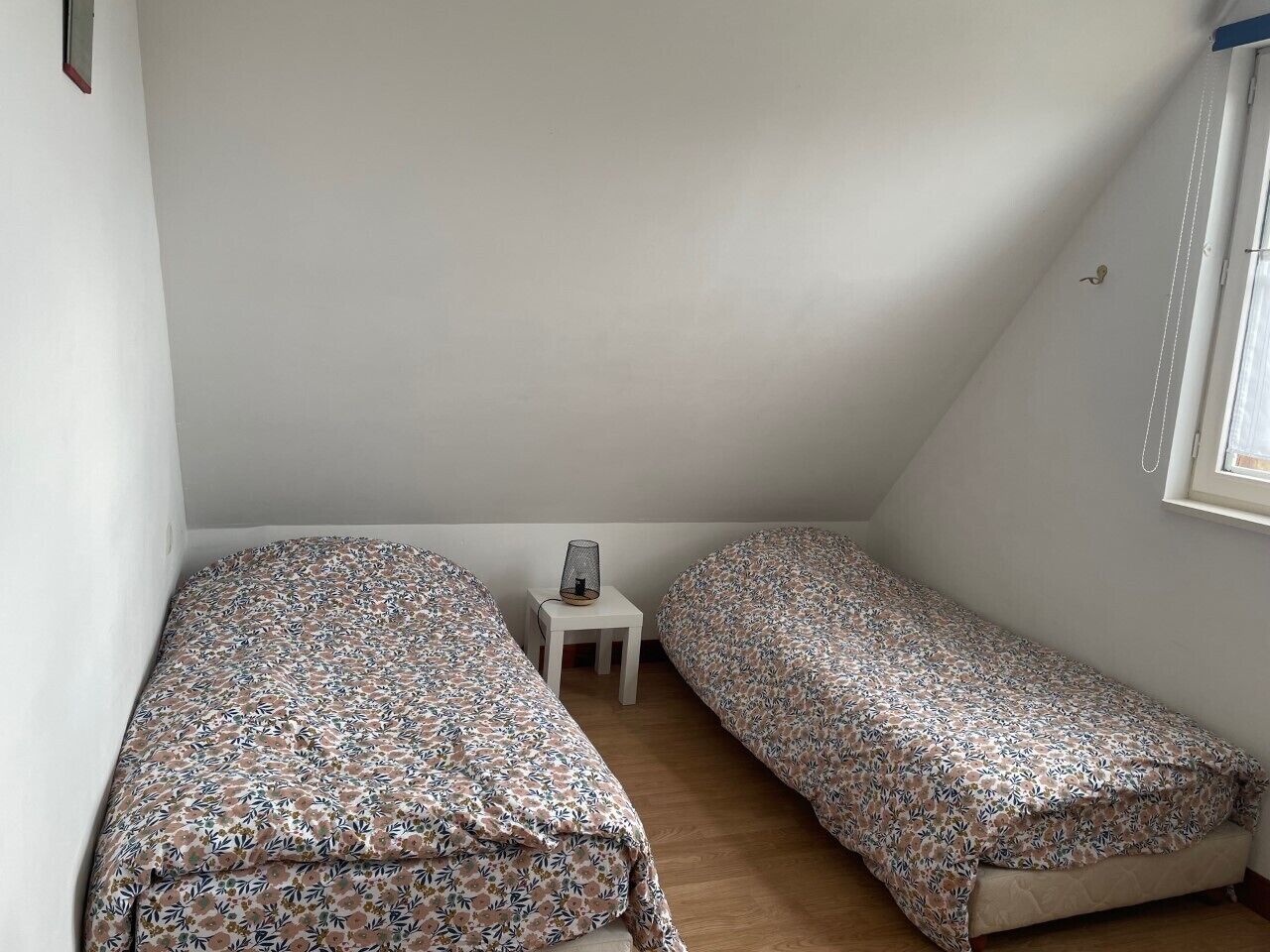 Quarto sótão pequeno e triangular com duas camas de solteiro, uma mesinha e uma janela.