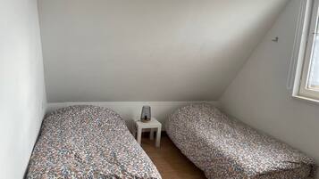 Quarto sótão pequeno e triangular com duas camas de solteiro, uma mesinha e uma janela.