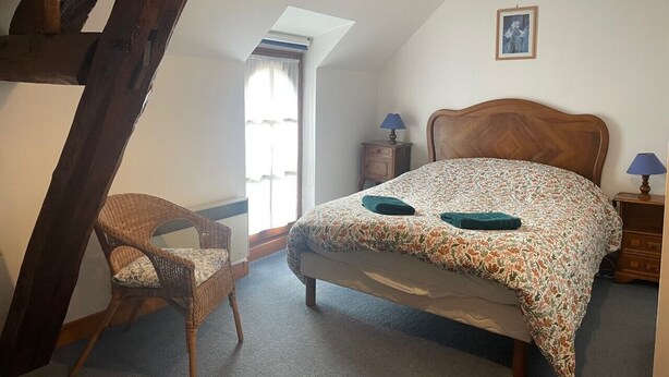 Un dormitorio con techo inclinado, una cama con colcha estampada, una silla de mimbre, una mesita de noche con lámpara y un cuadro enmarcado en la pared.