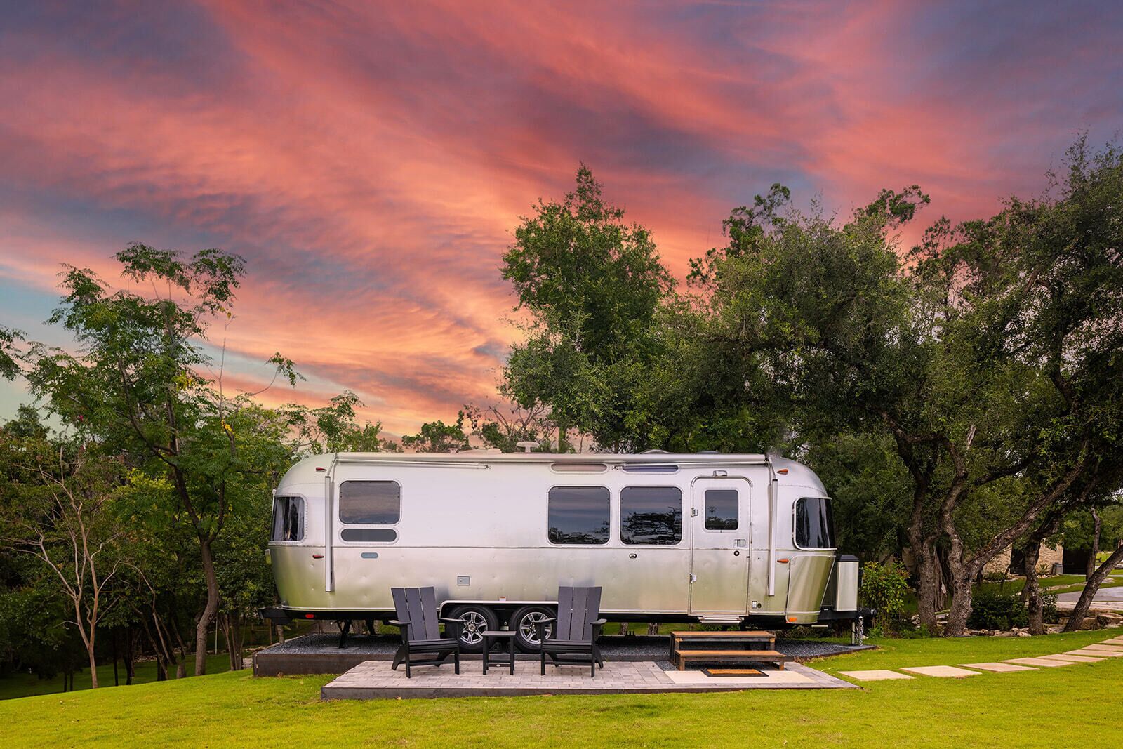 A stationary RV with a patio area, including a table and chairs, surrounded by trees and a grassy lawn.