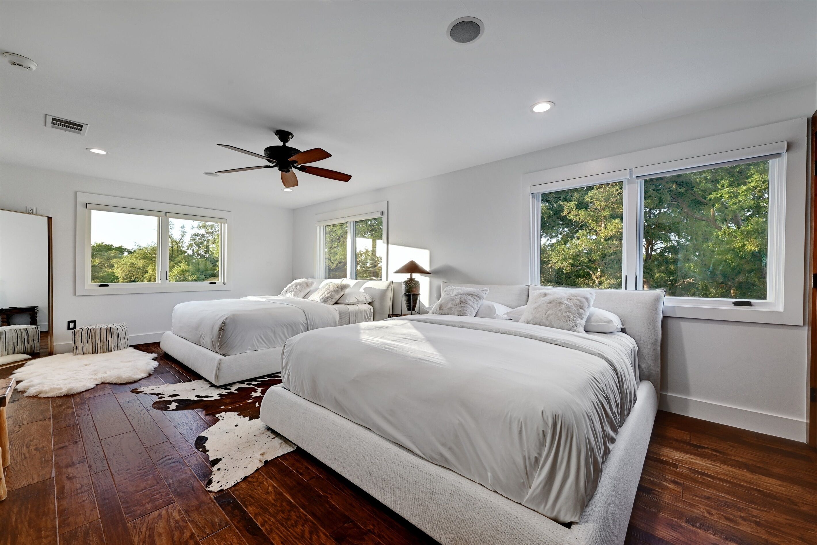 A bedroom with two large beds, a ceiling fan, and large windows offering a view of trees.