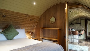 A cozy bedroom with a wooden ceiling, a bed with a green pillow, a bedside table with a lamp, and a view into a kitchen area with bar stools. - Hare's View Underground House (Rhayader)