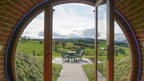 A circular brick archway leading to an outdoor patio with a table and chairs, surrounded by a grassy landscape.