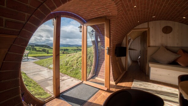 A circular room with a brick archway leading to a glass door, a wooden bench outside, and a bed with pillows inside.