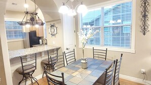 A dining area with a large table, six chairs, and a chandelier. There is a kitchen visible through an archway, and a window with blinds. - Paradise Cove Walk-in Log Cabin by Silver Dollar City & Table Rock Lake! (Branson)