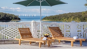 Een terras met houten lounge stoelen en een parasol, met uitzicht op een waterpartij met zeilboten.