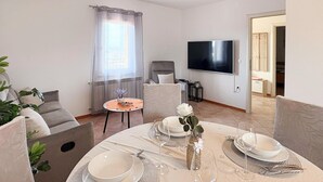 Een eetruimte met een ronde tafel voor vier personen, een flatscreen televisie aan de muur bevestigd en een raam met transparante gordijnen.