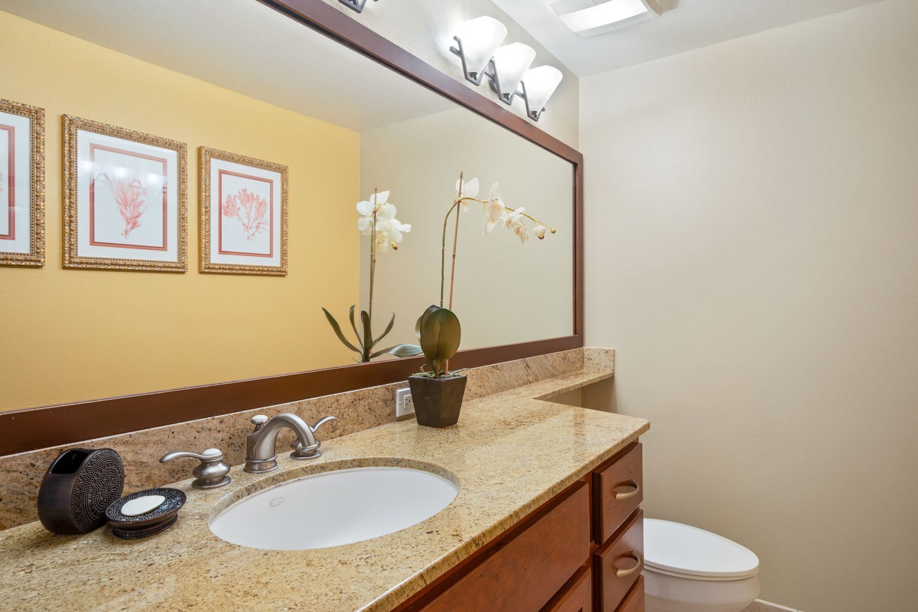 A bathroom with a granite countertop, a white sink, a wooden vanity, a toilet, and framed artwork on the wall.