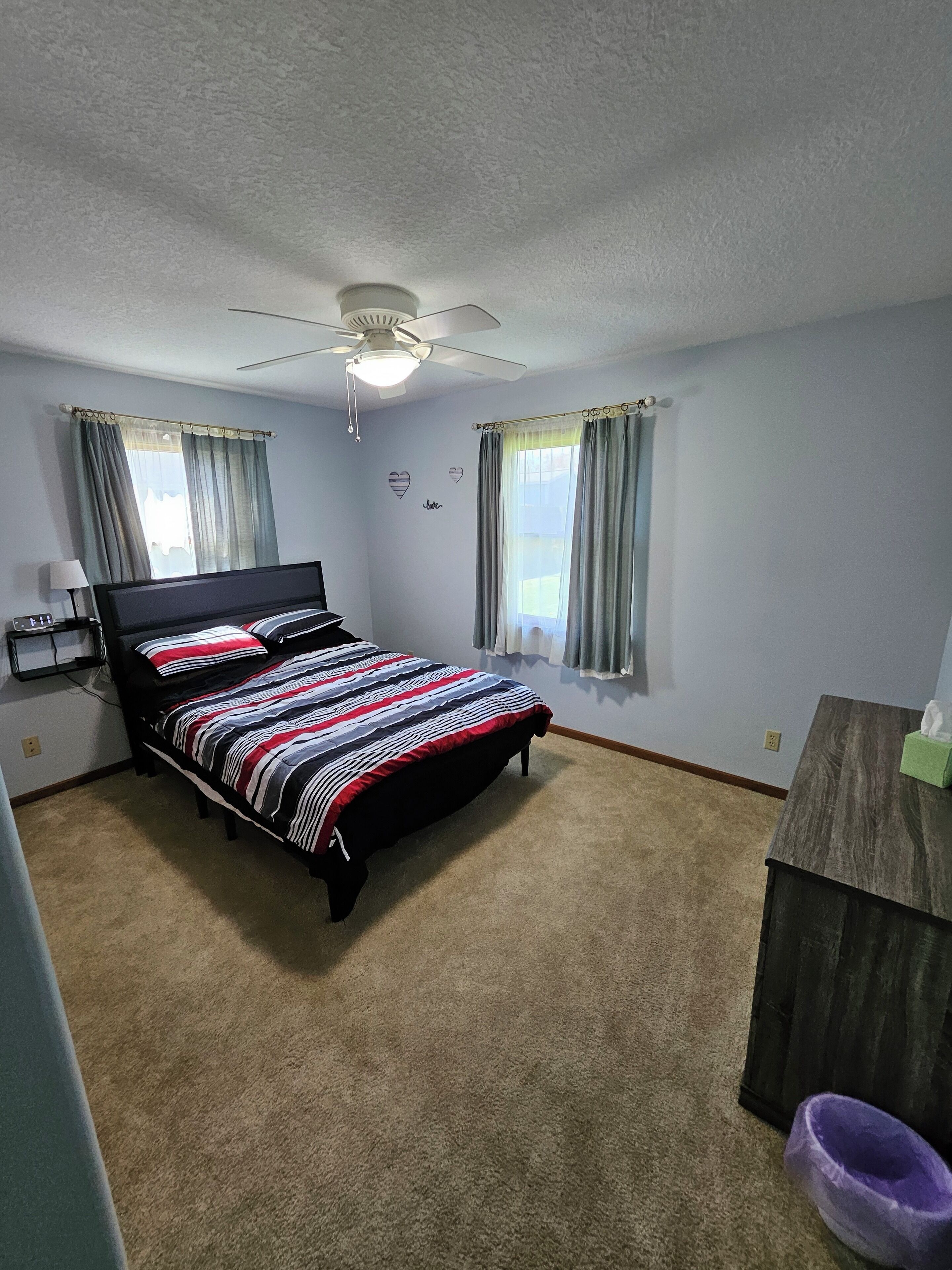 Quarto com cama, ventoinha no teto, duas janelas com cortinas, mesa de cabeceira e cômoda.