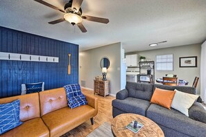 Ein Wohnzimmer mit braunem Ledersofa, einem grauen Sofa und einem runden Couchtisch. Es gibt einen Deckenventilator, eine Wandleuchte und im Hintergrund eine Küchenzeile.