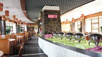 A restaurant with round tables and chairs, a buffet area with food containers, and a stone pillar with a sign.