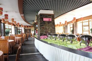 Un restaurante con mesas y sillas redondas, una zona de bufé con recipientes de comida y un pilar de piedra con un cartel.