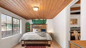 Een slaapkamer met een houten plafond, een bed met hoofdbord, een nachtkastje, een stoel en een raam met jaloezieën.
