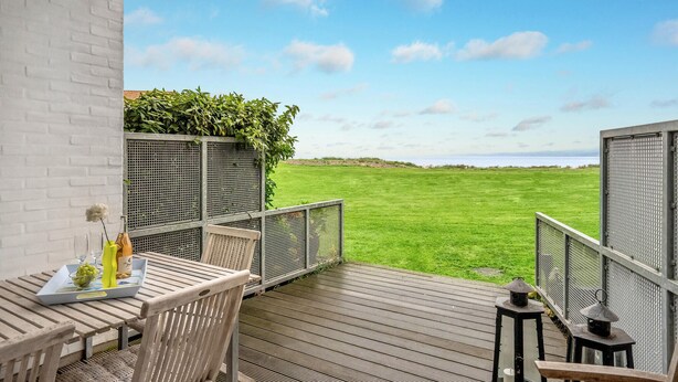 Eine Holzterrasse mit Tisch und Stühlen, die auf eine Wiese und das Meer blickt.