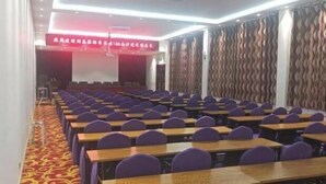 A conference room with rows of tables and chairs, a stage with a red banner, and patterned curtains.
