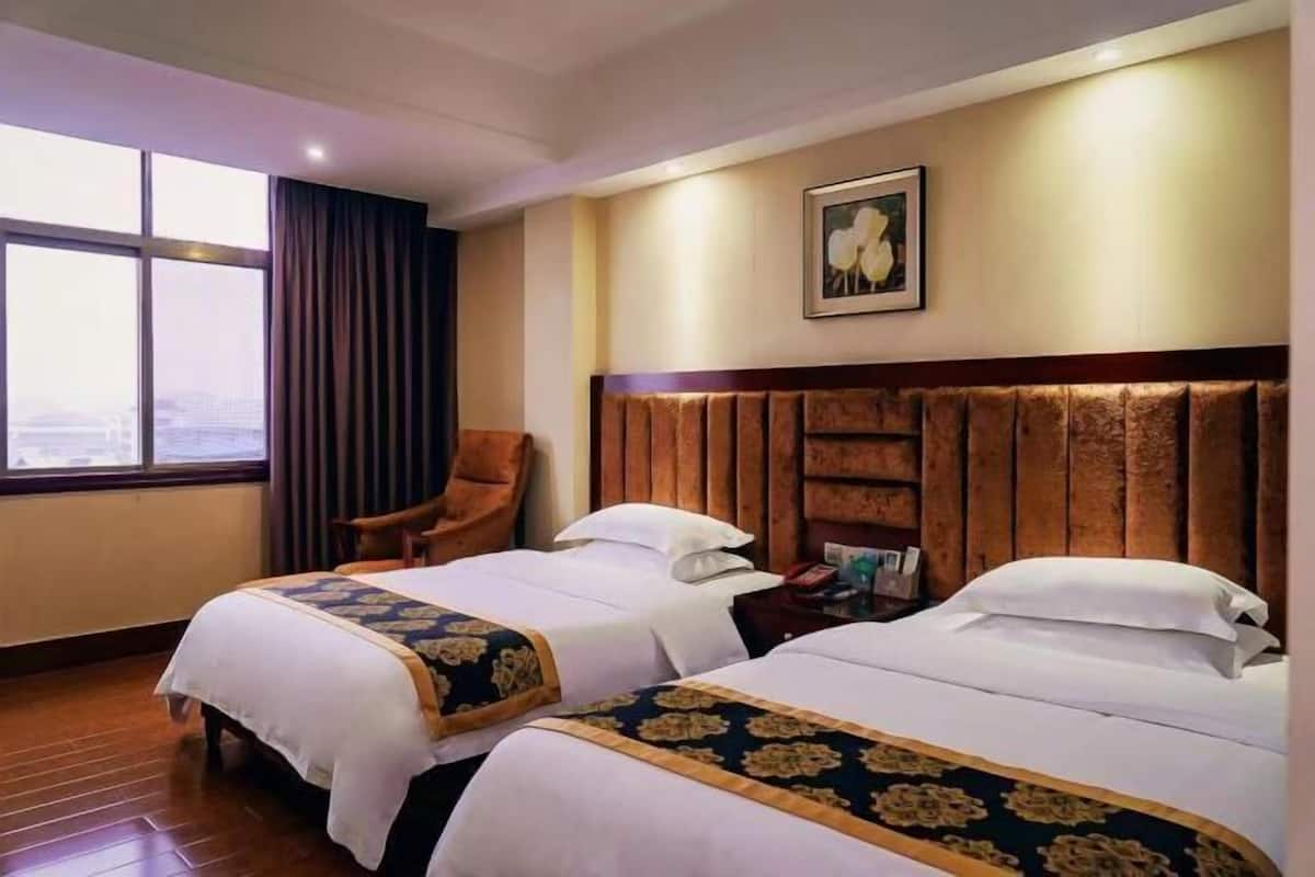 Habitación de hotel con dos camas, cabecera de madera, una silla, ventana con cortinas e imagen enmarcada en la pared.