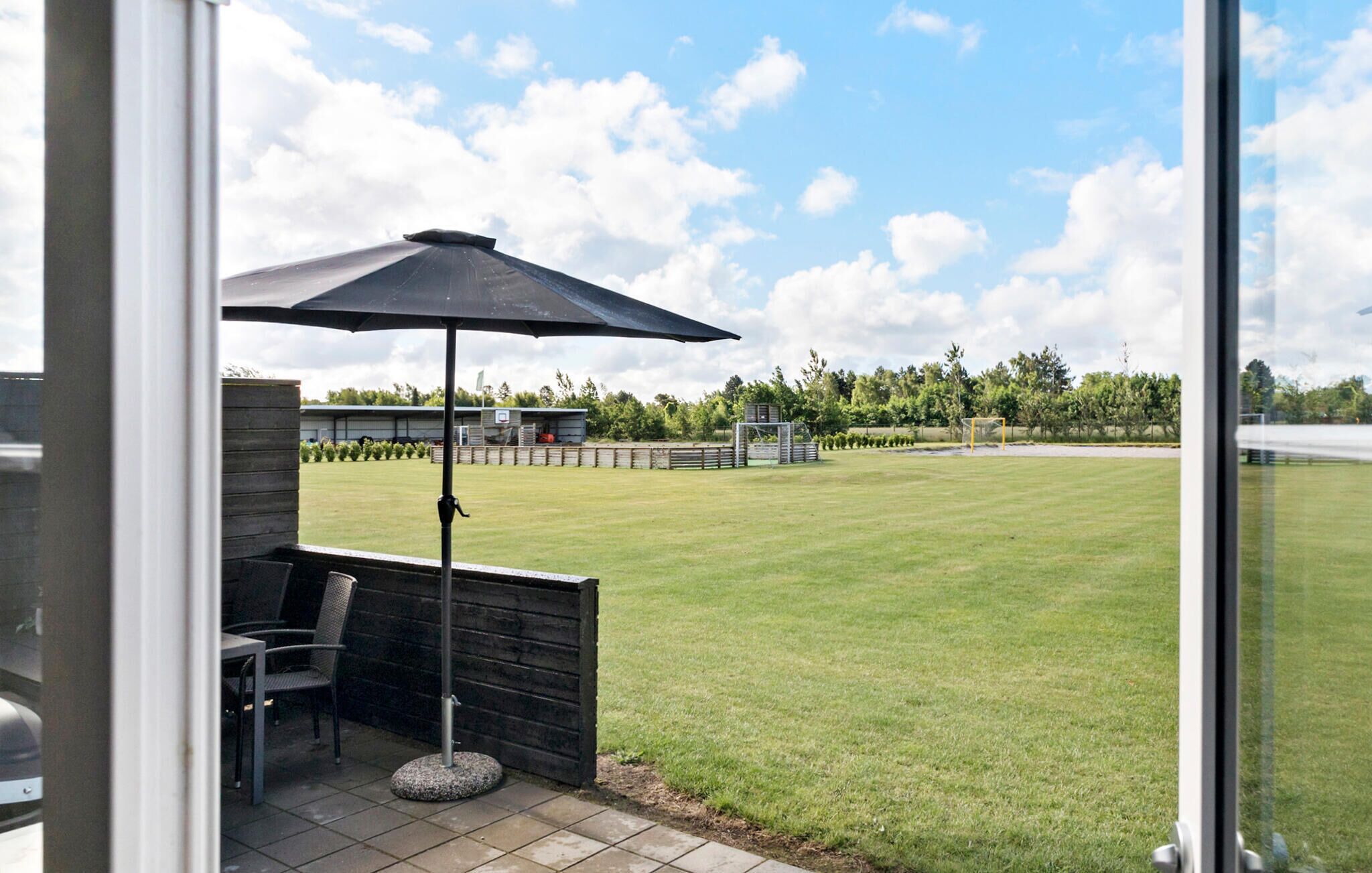 A patio area with a table and chairs, a large umbrella, and a view of a grassy field with a fence and trees in the background.
