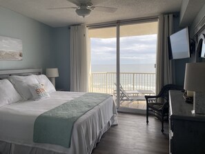 Ein Hotelzimmer mit Bett, Schreibtisch, Stuhl, Fernseher und einem Balkon mit Blick auf den Strand.