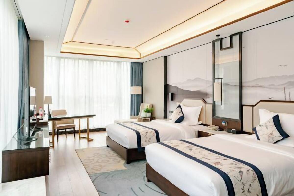 A hotel room with two beds, a desk, a chair, a desk lamp, a nightstand, and a large window with curtains.