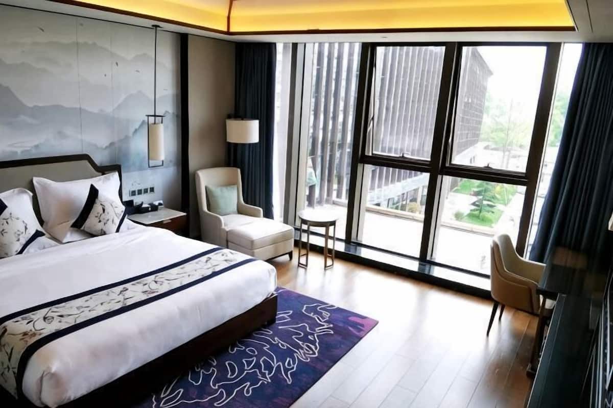 A modern hotel room with a large bed, a desk, a chair, and a view of the outside through large windows.