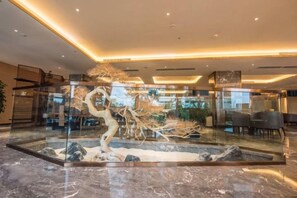 A lobby with a glass enclosure featuring a sculptural tree and rocks, marble flooring, and modern lighting.