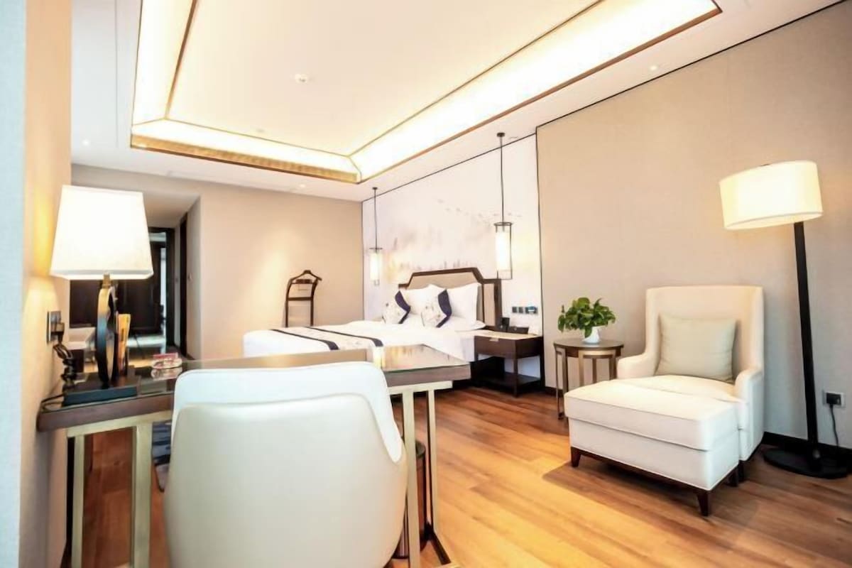 A modern hotel room with a bed, a desk with a lamp, a chair, a side table with a plant, and a floor lamp.