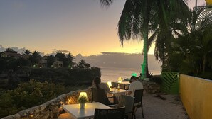 Un área de comedor al aire libre con mesas y sillas, con vista al mar durante el atardecer.