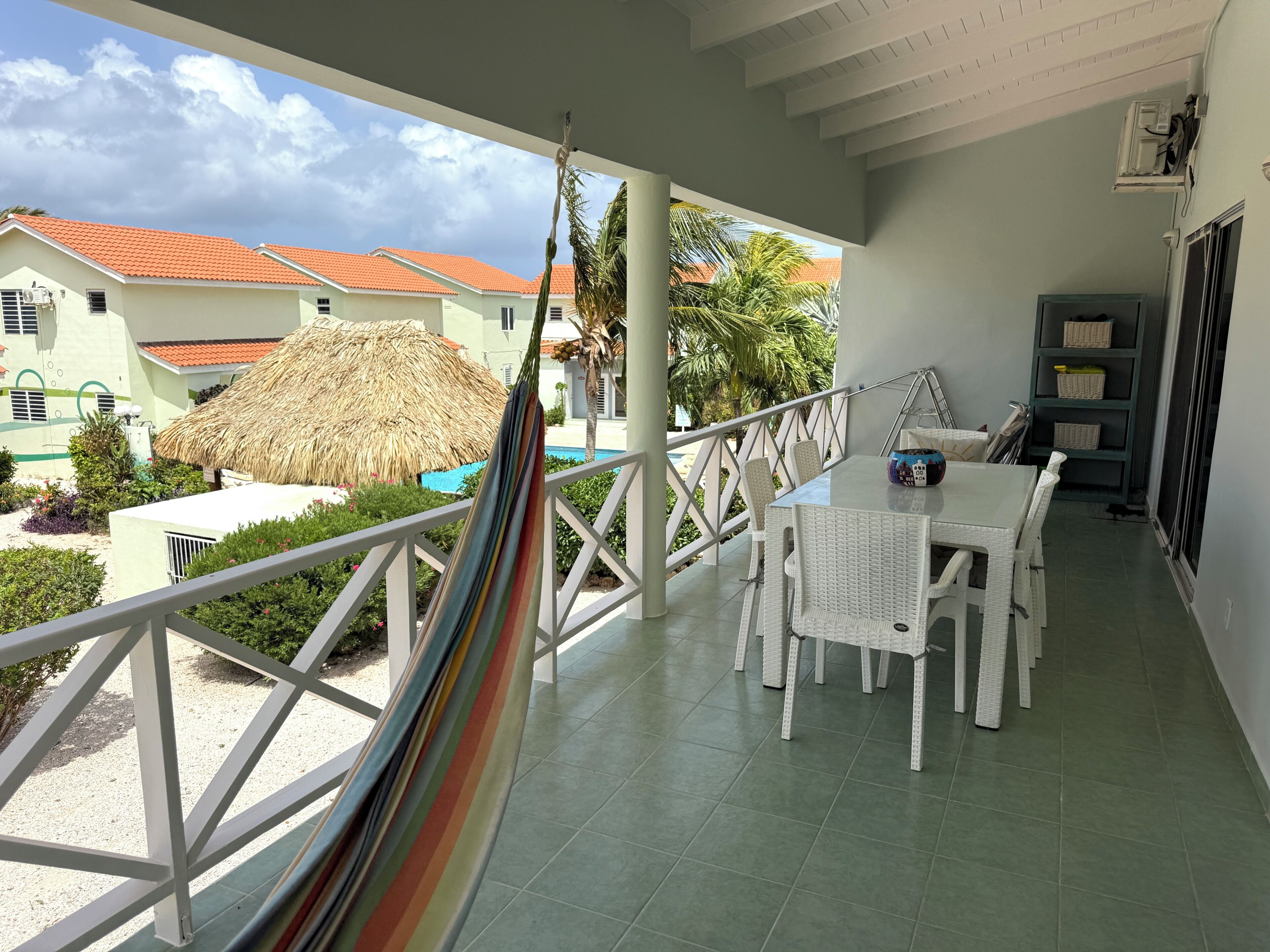 A balcony with a white table and chairs, a hammock, a pool, and a view of buildings with red roofs.