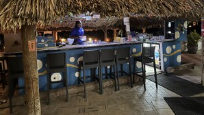 Una zona de bar con techo de paja, taburetes y una barra con diferentes objetos.