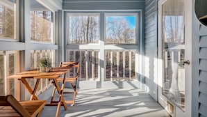 A covered porch with a wooden table and chairs, large windows, and a door.