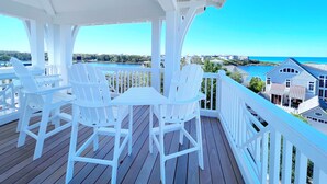 Eine überdachte Terrasse im Freien mit weißen Stühlen und einem Tisch, mit Blick auf ein Gewässer und Gebäude.