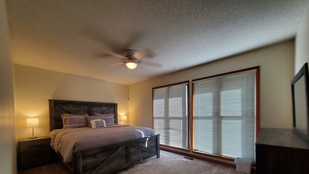 A bedroom with a bed, nightstands, a ceiling fan, and large windows with blinds. - Comfy Newly Furnished Home (Warrensburg)