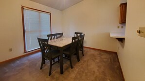 A room with a dining table and four chairs, a ceiling fan, a window with blinds, and a kitchen area with cabinets. - Comfy Newly Furnished Home (Warrensburg)