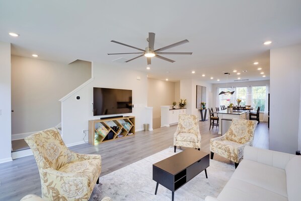 A spacious living room with a modern ceiling fan, a flat-screen TV, and a dining area with a kitchen in the background. - Durham Vacation Rental w/ Community Pools! (Durham)