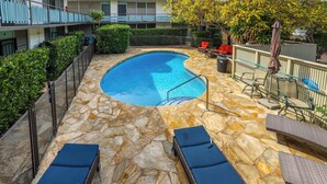 Piscina con suelo de piedra, sillones de descanso y un área vallada con un árbol y mobiliario exterior.