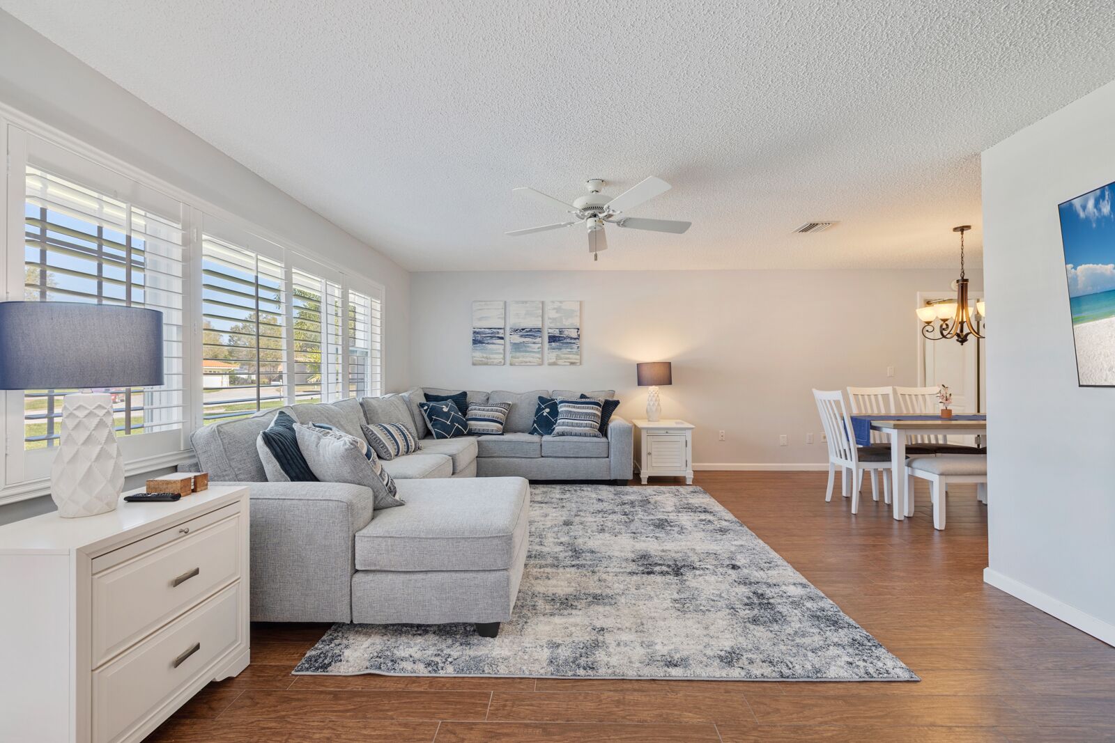 A modern living room with a grey sectional sofa, a dining area with white furniture, and a ceiling fan.