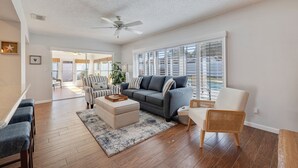 A spacious living room with a gray sofa, striped armchair, and a white coffee table. A ceiling fan and large windows provide natural light. - My Happy Place – Heated Pool, Dual Living Rooms, Screened Lanai & Nearby Indian Rocks Beach (Seminole)