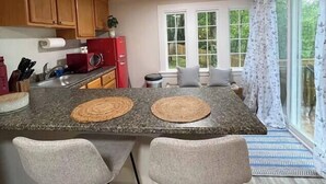 A kitchen with a large island, two bar stools, wooden cabinets, a red refrigerator, and a view of the outdoors through large windows. - Spacious 1 Bedroom Cottage Close to Everything 5 (Edgecomb)