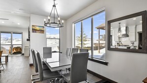 Un espace repas moderne avec une grande table et des chaises, de grandes fenêtres offrant une vue sur le paysage enneigé, et une cuisine visible dans le reflet.