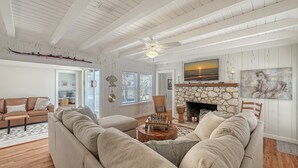 A spacious living room with a high ceiling, a stone fireplace, and a large window with blinds.